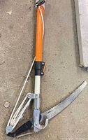 Close view of cutting head with curved saw blade and lopper mechanism on orange fiberglass pole