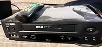 Front three-quarter view of RCA VCR with remote resting on top showing branding, buttons and AV input jacks
