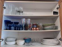 Cabinet with assorted dishes: Corelle plates/bowls, blue goblets, iridescent glasses, copper mugs, Coca-Cola glasses, Barcela bowls.