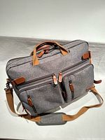 Front view showing gray canvas body, dual front zip pockets, tan faux leather trim, detachable shoulder strap attached