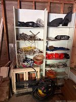 Overall view: two white shelving units filled with rope coils, reels, plastic parts, vent grates, wooden crate with copper pipe, Shop-Vac on floor
