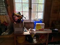 Bench area showing box with plastic toys, lanterns, figurine, blue crate with rope, repair kit box, extension cords below
