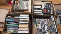 Four boxes filled with assorted hardcover and paperback books including visible Clive Cussler, Walter Cronkite, Net Force titles