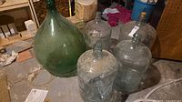 Group of one green demijohn and four clear carboys on floor