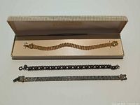 Three bracelets displayed: gold-tone in case, black heart link, silver panel link