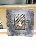 Front view of square ceramic wall sculpture showing purple glaze, metal lattice, hanging teardrop element