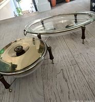 Oval glass casserole with glass lid and round casserole with brass lid on respective brass-tone cradles