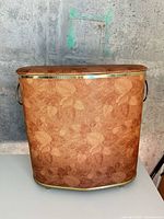 Front view of oval hamper showing leaf pattern vinyl exterior, gold-tone trim and handles