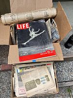 Open box showing LIFE magazine March 26, 1953 issue atop stack of newspapers and rolled paper