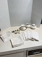 Group shot of napkins, porcelain cup, saucer, creamer, sugar bowl and spoons