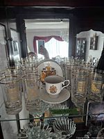 cabinet shelf with multiple gold-crest tumblers, one porcelain cup and saucer visible