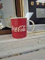 Red ceramic Coca-Cola mug in front of framed sign and pin display