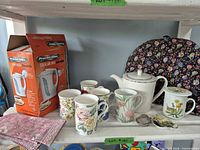 all items on shelf: six mugs, teapot, infuser, boxed kettle, tea cozy visible