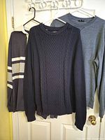 Three men’s tops hanging: navy cable-knit sweater, heather blue sweatshirt, navy/white striped rugby shirt