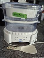 Front view of Oster food steamer with two clear baskets, white base, lid and floral spoon