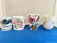 Front view of four mugs: Lazzaro logo, insect quote, books graphic, blue bird