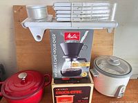 All five items grouped: Melitta pour-over set box, Rival Crock-ette box, Zojirushi rice cooker, red Dutch oven, mandoline slicer with attachments