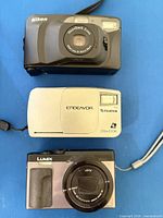 Front view of three compact cameras showing models, lenses, flash units and wrist straps