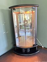 Front view showing curved glass door, mirrored back, interior light on, black matte frame and crescent base