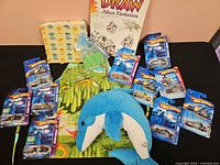 Group shot of all lot items: nine carded Hot Wheels vehicles, dinosaur apron set, dolphin plush, drawing book, bead pouch