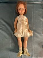 Full view of one composition doll lying flat wearing satin dress, socks, shoes