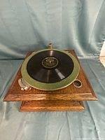 Three-quarter view of phonograph base showing oak cabinet, green felt platter, record, crank, and horn mount opening