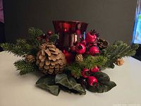 Front view of centerpiece showing red glass holder, pine cones, berries, greenery
