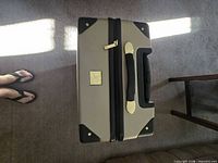Top view of closed suitcase showing beige shell, black trim, carry handle and gold-tone logo plaque