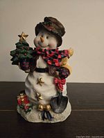 Front view of snowman figurine showing tree, shovel, gifts, bells, attire