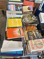 Tabletop view showing multiple stacks of hardcover and paperback books of varying genres, dust jackets visible, some with sticky notes