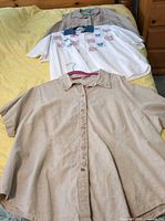 Three plus size shirts: two beige, one white with embroidered hearts, laid out on bed