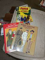 Stack of four books: The Phantom comic, unidentified comic partially visible, John Wayne and Santa Claus paper doll books