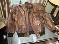 Front view of brown leather bomber jacket laid flat showing pockets, zipper, collar, cuffs, waistband