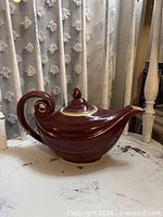 Left side view of maroon teapot showing curled handle, spout, lid, gold accents