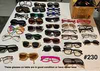Overview of table with approximately 30 pairs assorted eyeglasses and sunglasses, box with additional pieces