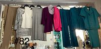 Nine clothing pieces hanging on rack: beige pants, striped top, white sleeveless top, grey sleeveless dress, plum top, checkered shirt, pink cardigan, turquoise pants and top