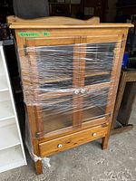 Front view of pine cabinet wrapped in plastic showing glass doors and bottom drawer