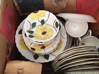 Stack of plates and cups in box showing yellow flower and green leaf design on white ceramic dishware.