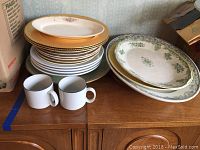 Stack of multiple plates including stoneware plates with gold rims, green floral design plates, two white ceramic mugs, and gold chargers.