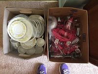 Two cardboard boxes filled with Nikko dishes with a yellow floral pattern on the left box including saucers, plates, and cups. The right box contains assorted glassware in various sizes wrapped in bubble wrap and pink cloth.