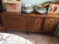 Full view of the wooden buffet sideboard with several plates, mugs and boxes on top, showing front drawers and cabinet doors and decorative molding.