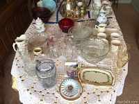 Wide shot of table exhibiting multiple glass, crystal, and ceramic items including pitchers, bowls, ashtrays, trays, and glasses on a crocheted tablecloth.