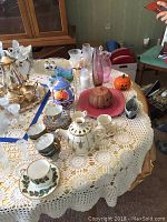 Photo of table with assorted glassware, Pyrex bowls, porcelain tea cups with saucers, and decorative glass items on a crocheted tablecloth