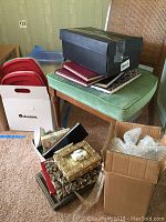 Photo showing a collection of items including books stacked on a green padded stool, photo albums, decorative photo frames, a black shoebox, and two cardboard boxes containing various items including bubble wrap and red trays.