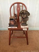 Front view of small wooden chair with crack on backrest, metal bust of a child, and metal mailbox coin box, all placed on the chair seat.