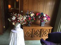 Wide view of three faux flower arrangements displayed on wooden console and small pedestal table with tablecloth