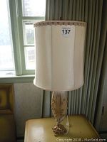 Full view of a tan floral ceramic table lamp with fabric shade, showing the lamp standing on a small table near window.