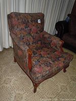Front and slight angle view of armchair showing dark floral upholstered seat, back, and armrests. Carved wooden arms and feet are visible, fabric appears faded and soiled in areas.