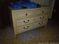 Full front view of the cream-colored 3 drawer wood dresser with floral carvings on the top drawer and metal handles on the bottom drawers.