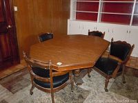 Photo of wood dining table with 4 matching captain's chairs, showing overall set in room with carpet and wood-paneled wall.
